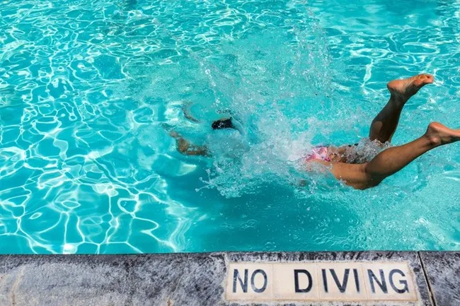Child diving into a clear blue pool near a concrete ‘No Diving’ warning sign