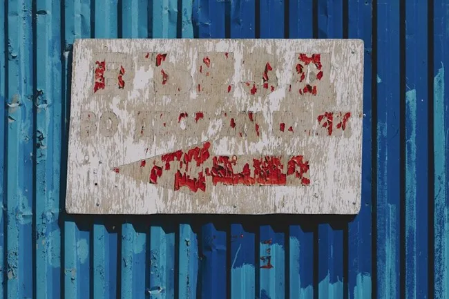 Faded and peeling wooden sign on a blue corrugated metal wall, showing wear from outdoor exposure