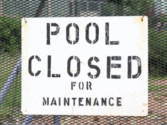 Weathered ‘Pool Closed for Maintenance’ sign on a black metal fence at an outdoor swimming area