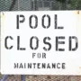Weathered ‘Pool Closed for Maintenance’ sign on a black metal fence at an outdoor swimming area