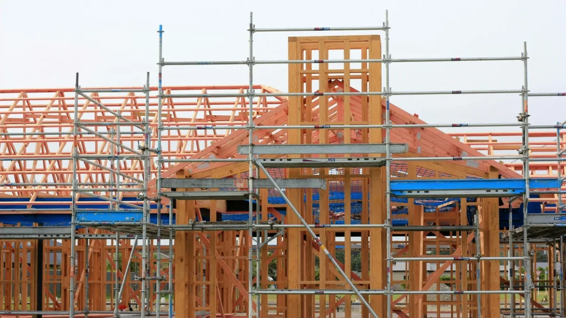 Timber frame home under construction with exposed posts, beams, and roof trusses surrounded by steel scaffolding.