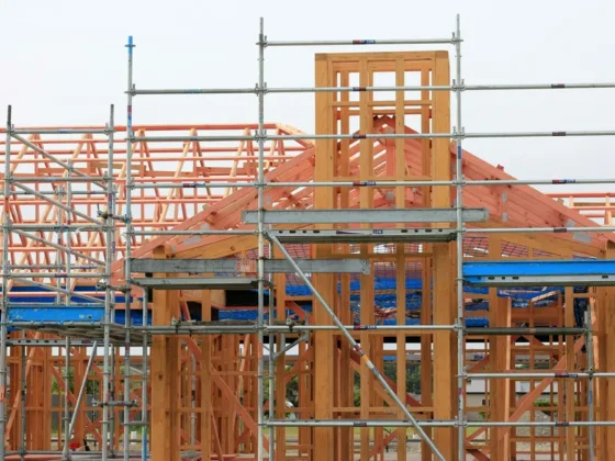 Timber frame home under construction with exposed posts, beams, and roof trusses surrounded by steel scaffolding.