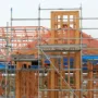 Timber frame home under construction with exposed posts, beams, and roof trusses surrounded by steel scaffolding.